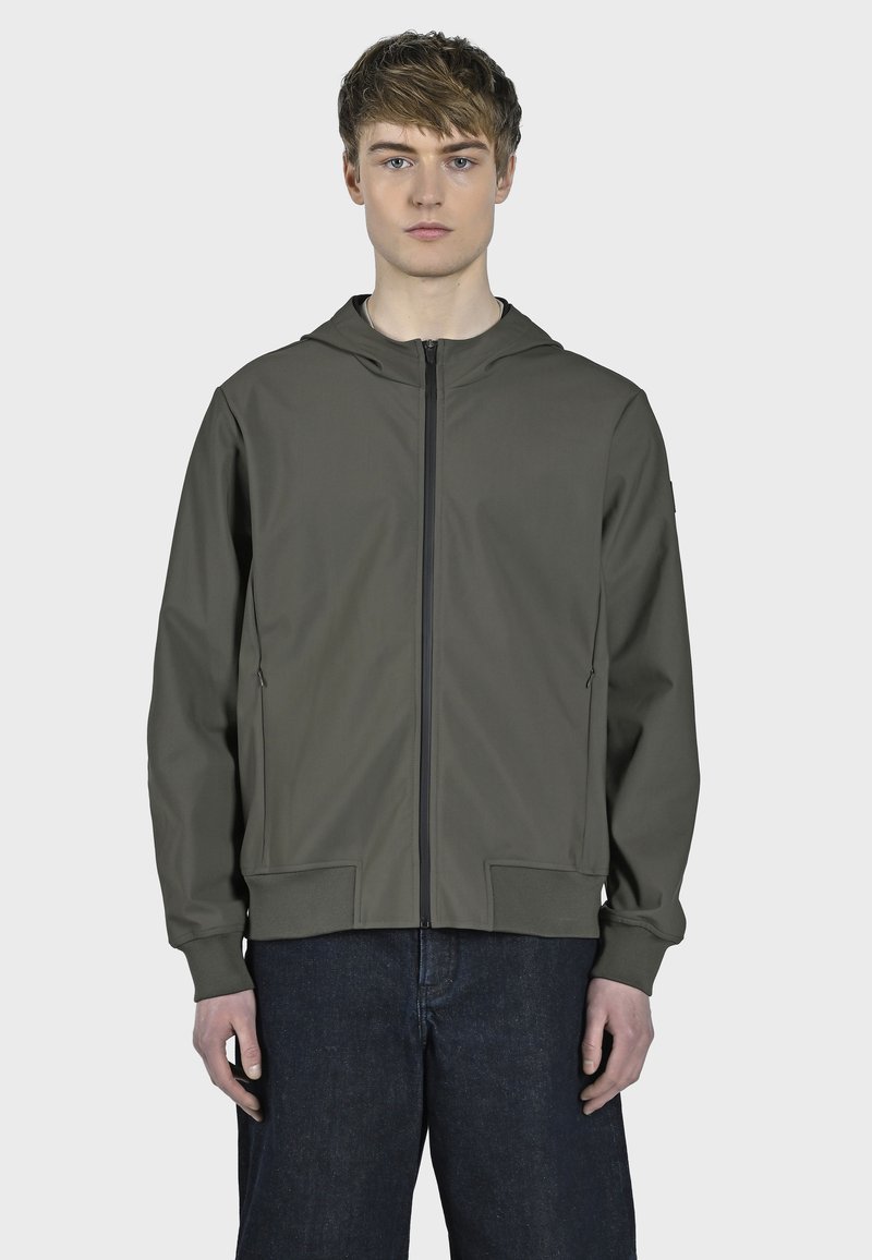 Giacca con cappuccio verde oliva, tessuto leggero, chiusura con zip, due tasche frontali, polsini e orlo a costine. Il modello si trova davanti a uno sfondo neutro.