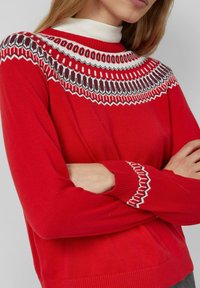 Czerwony dziergany sweter z okrągłym dekoltem oraz dekoracyjnymi wzorami w białym i ciemnym odcieniu wokół ramion i mankietów.