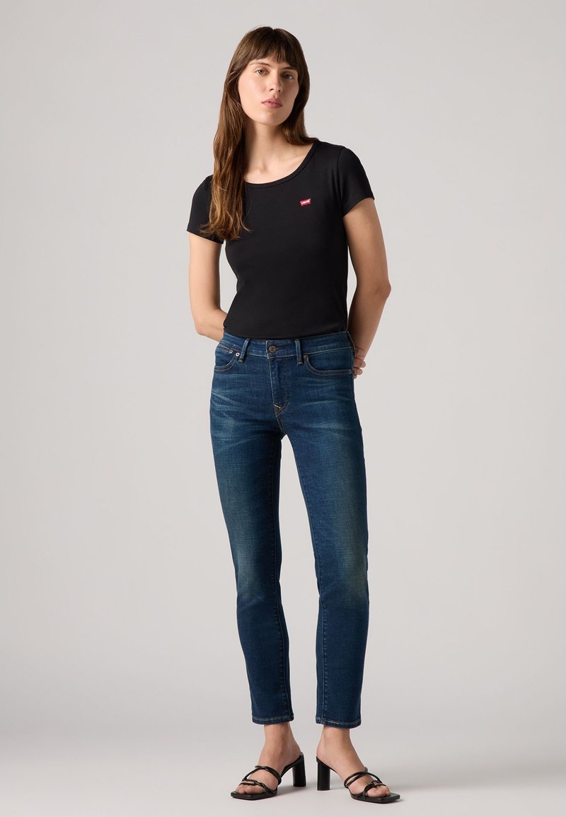 Schwarzes T-Shirt mit kurzen Ärmeln und kleinem roten Logo, kombiniert mit blauen Skinny-Jeans und schwarzen High Heels. Schlichte, figurbetonte Silhouette.
