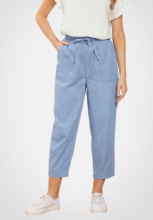 Kobieta ubraną w błękitne, krótkie spodnie z przodu z kieszeniami i białe trampki, stojąca z jedną ręką w kieszeni.