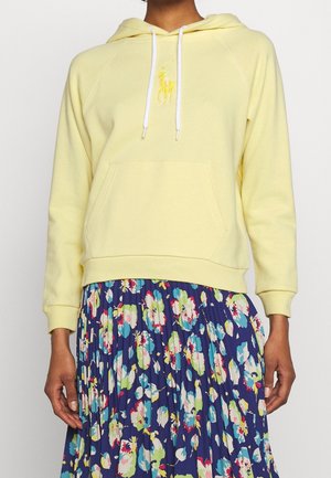 Person wearing a pale yellow hoodie with kangaroo pocket and white drawstrings, paired with a pleated navy skirt featuring colorful floral patterns.