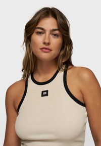 Top sin mangas de canalé en beige con un escote negro. Presenta un pequeño parche de logo negro en el centro. El material parece elástico y suave.