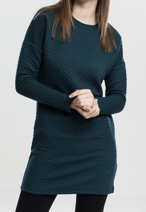 Vrouw die een donkergroene gegolfde lange mouwen jurk draagt, staand met haar handen gevouwen voor zich tegen een effen achtergrond.