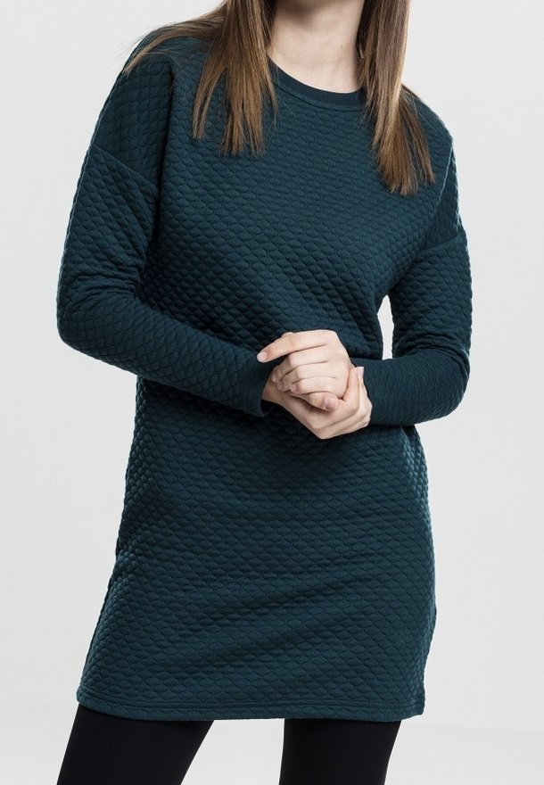 Femme portant une robe longue à manches longues texturée bleu sarcelle foncé, debout, les mains jointes devant elle sur un fond uni.