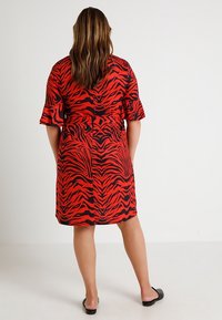 Vestido con estampado de tigre en rojo y negro, con mangas cortas y volantes, cintura ceñida, corte a la altura de la rodilla y textura de tela suave. Sandalias negras.