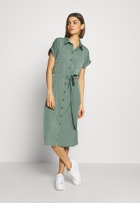 Abito camicia verde a maniche corte con collo a camicia, apertura a bottoni frontale, cintura da annodare in vita e spacco laterale; abbinato a sneaker bianche.