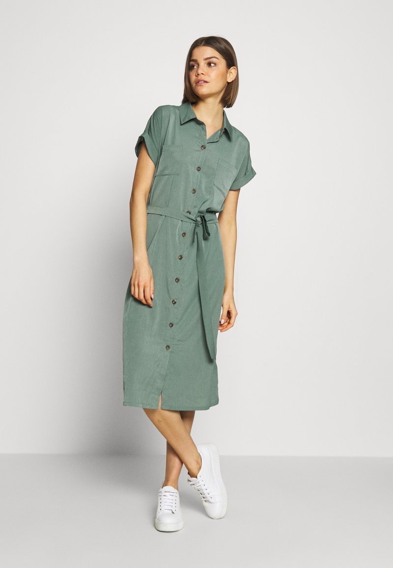 Abito camicia verde a maniche corte con collo a camicia, apertura a bottoni frontale, cintura da annodare in vita e spacco laterale; abbinato a sneaker bianche.
