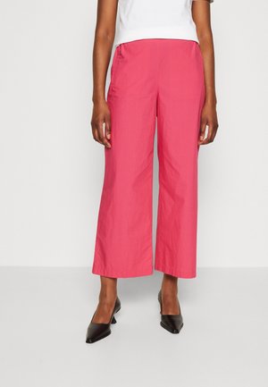 Vrouw draagt een roze broek met hoge taille en wijde pijpen tot net boven de enkels, een wit topje en zwarte pumps met puntige neus, staand tegen een effen lichte achtergrond.