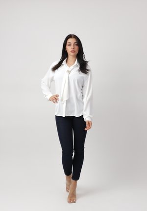 Mujer con cabello largo y oscuro, vistiendo una blusa blanca con detalle de corbata en el cuello, jeans oscuros ajustados y sandalias de tacón alto color nude, posando con confianza.