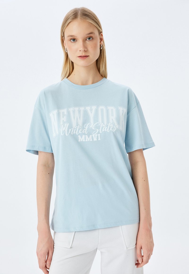 Lichtblauwe katoenen t-shirt met korte mouwen, met op de voorkant de witte tekst "NEW YORK Verenigde Staten MMVII" bedrukt.
