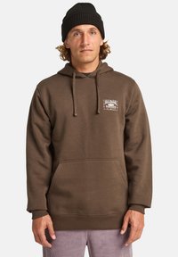 Sweat à capuche marron en tissu doux, avec une poche kangourou et un cordon de serrage pour la capuche. Détail du logo sur la poitrine gauche. Manches à bord côtelé.