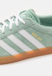 Zelený semišový teniska s třemi bílými pruhy, zlatým nápisem "GAZELLE" a texturovanou podrážkou barvy gumy. Má bílé tkaničky a polstrovaný límec.