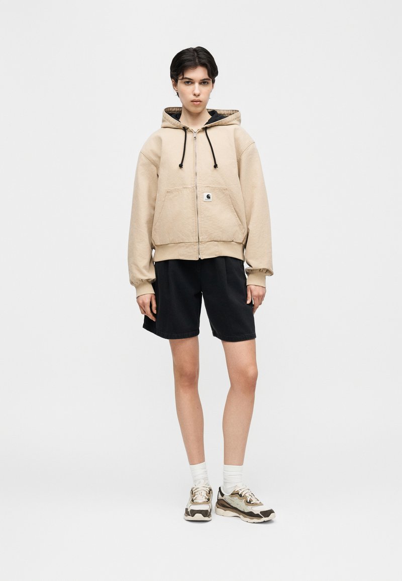 Jeune personne debout, portant un sweat à capuche beige zippé, un short noir, des chaussettes blanches et des baskets, sur un fond clair uni.