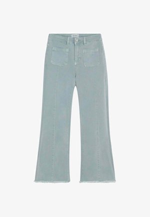 Mujer joven de pie con sudadera gris oscuro, jeans acampanados cortos azul claro y zapatillas blancas, mirando hacia un lado.