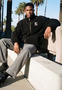 Mladý muž sedící na venkovní betonové lavičce pod palmami, oblečený v černé fleecové bundě se zipem, šedých džínách a černých teniskách.