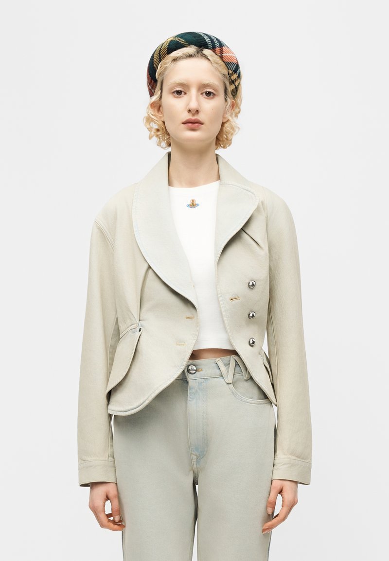 Νεαρή γυναίκα με ξανθά σγουρά μαλλιά φοράει μια κορδέλα με σχέδια, μπεζ cropped σακάκι με ασημένια κουμπιά, λευκή μπλούζα και ανοιχτόχρωμο τζιν.