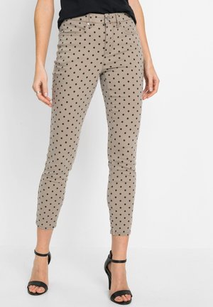 Pantaloni beige a taglio aderente con pois neri, indossati con décolleté nere con cinturino alla caviglia e top nero.
