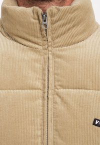 Beige corduroy jack met een hoge kraag, voorrits en zwart logo-hangar. Kenmerkt zich door een gequiltde textuur en verticale ribbelstructuur.