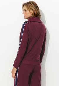 Veste de sport bordeaux avec un col, agrémentée de bandes latérales bleu marine et de poignets côtelés. Tissu lisse avec une coupe ajustée, associée à un pantalon assorti.