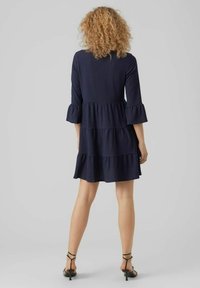 Dunkelblaues, gestuftes Kleid mit Drei-Viertel-Ärmeln und Rüschensäumen. Strukturierter Stoff mit fließender Silhouette, kombiniert mit schwarzen Riemchensandalen.