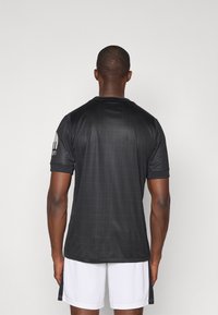 Maglietta sportiva nera con texture a rete e maniche corte, con un logo sulla manica sinistra. Indossata con pantaloni corti bianchi.