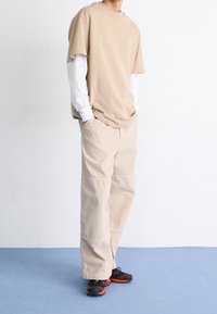 Camiseta oversized beige superpuesta sobre una camiseta de manga larga blanca, combinada con pantalones beige holgados y zapatillas deportivas negro-naranja.
