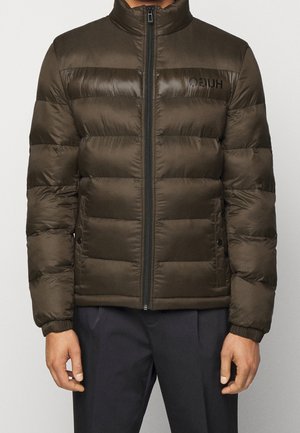 Homme portant une veste matelassée marron foncé avec une fermeture éclair frontale et un pantalon noir, debout les bras relâchés le long du corps.