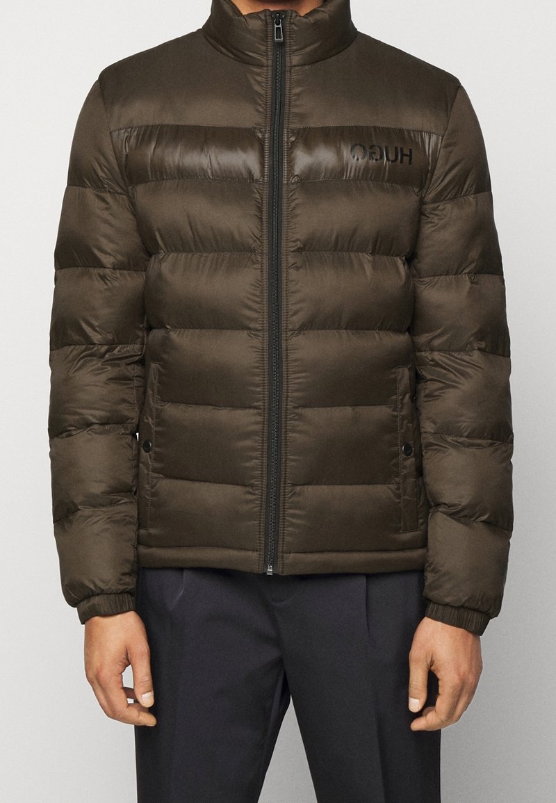 Homme portant une veste matelassée marron foncé avec une fermeture éclair frontale et un pantalon noir, debout les bras relâchés le long du corps.