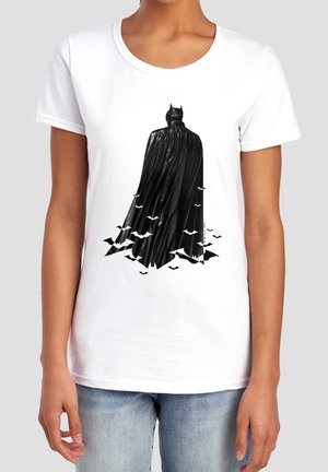 Black silhouette of Batman with flowing cape transforming into flying bats on white T-shirt worn by person in light blue jeans.