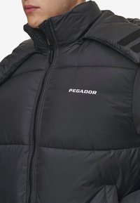 Black puffer jacket with a high collar, featuring a front zipper, horizontal stitching, and "PEGADOR" logo on the chest. Textured fabric.
