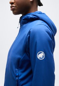 Άτομο που φοράει μπλε σακάκι με κουκούλα και λευκό λογότυπο μαμούθ στον επάνω μανίκι, κοιτάζοντας προς τα αριστερά σε ουδέτερο φόντο.