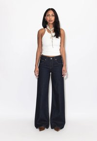 Kvinde med langt mørkt hår iført hvid ærmeløs top, mørke vidde jeans, brune spidse sko og et lyst tørklæde omkring halsen, stående.