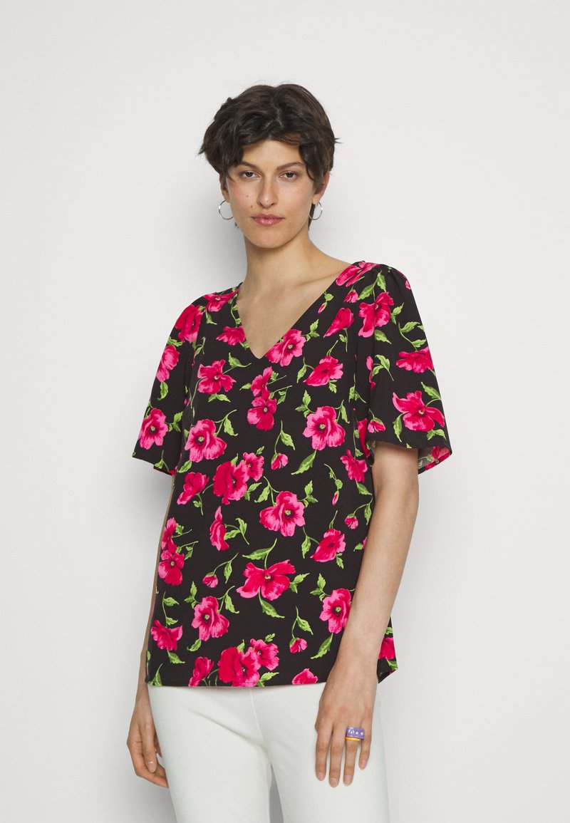 Blusa preta com padrões florais vibrantes em rosa e verde, mangas curtas e decote em V. Combinada com calças brancas e um anel.