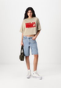 Beige cropped t-shirt med rød grafisk print, denim shorts, hvide højhælede sneakers og en grøn taske. Modellen står foran en hvid baggrund.