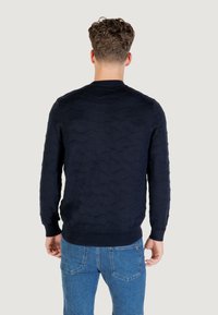 Dunkelblauer Pullover mit einem strukturierten Wellenmuster, Rundhalsausschnitt und gerippten Bündchen und Saum. Trägt eine helle Jeans, Ansicht von hinten.