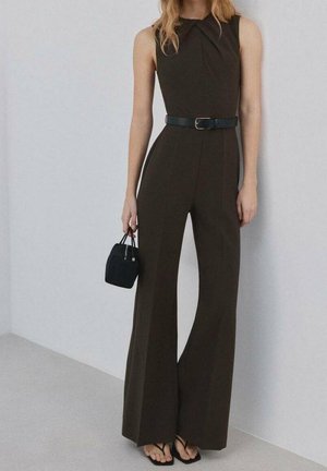 Kvinde iført en mørkebrun ærmeløs jumpsuit med sort bælte, der holder en lille sort håndtaske og bærer sorte sandaler.