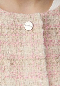 Beige textiel met een textuur en roze accenten, met een ronde zilveren speld met het "comma" logo, die een geweven ontwerp benadrukt.