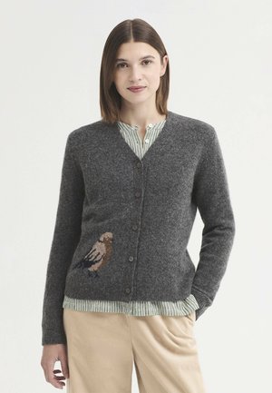 Jeune femme portant un cardigan gris boutonné avec un motif d'oiseau, une blouse rayée verte et un pantalon beige, debout avec une main dans la poche.