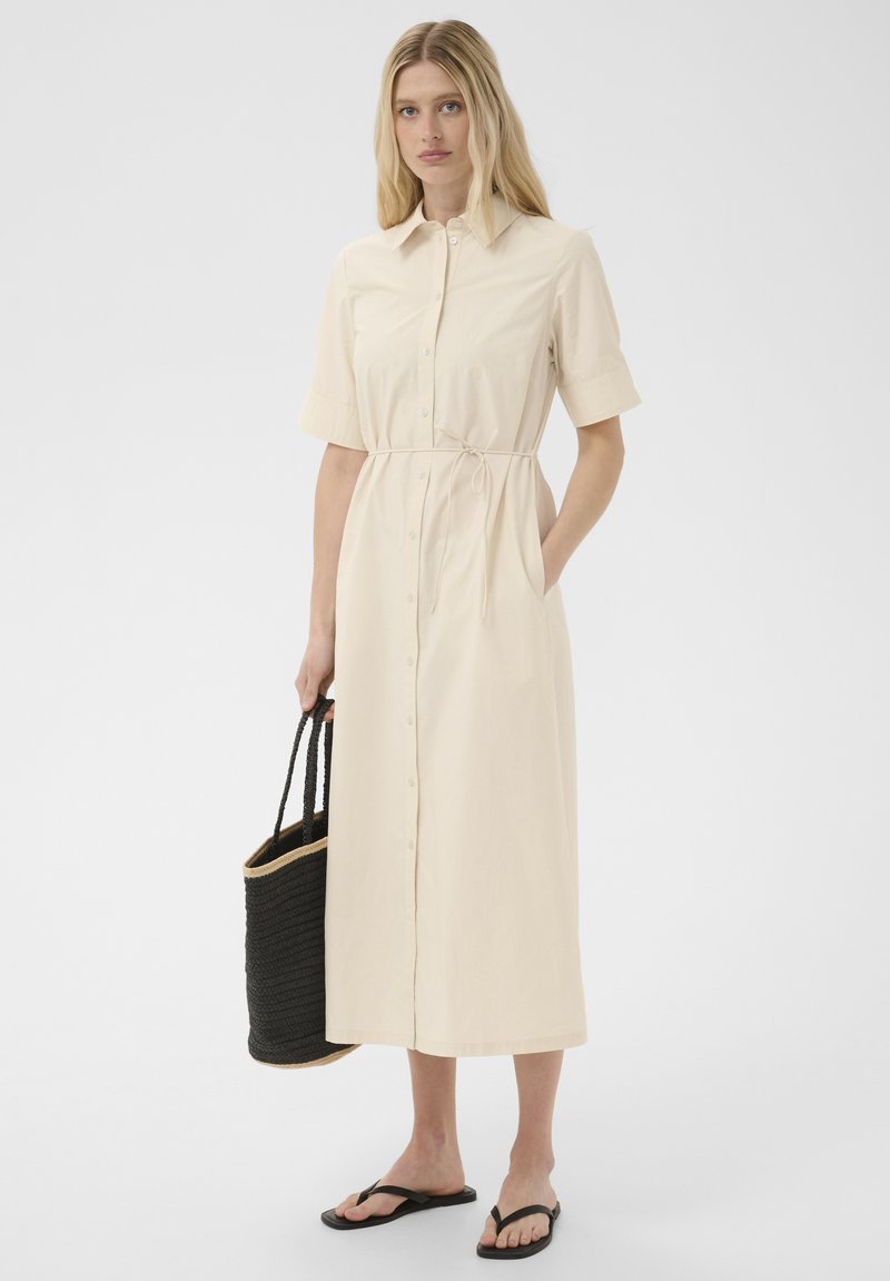 Femme portant une robe midi beige à boutons avec manches courtes, tenant un sac fourre-tout noir tissé, et portant des sandales tongs noires.