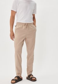 Beige lightweight trousers with elastic waistband and drawstring, featuring side pockets and a relaxed fit. Paired with brown slide sandals.