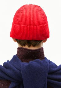 Persoon met de rug naar ons toe, gekleed in een rode gebreide beanie, een bessenrode gemêleerde coltrui en een blauwe trui die over hun schouders hangt.