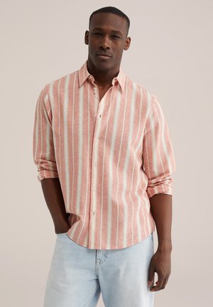 Mężczyzna w koszuli na guziki z pionowymi różowo-białymi paskami, z podwiniętymi rękawami, oraz jasnoniebieskich dżinsach, stojący na jednolitym tle.