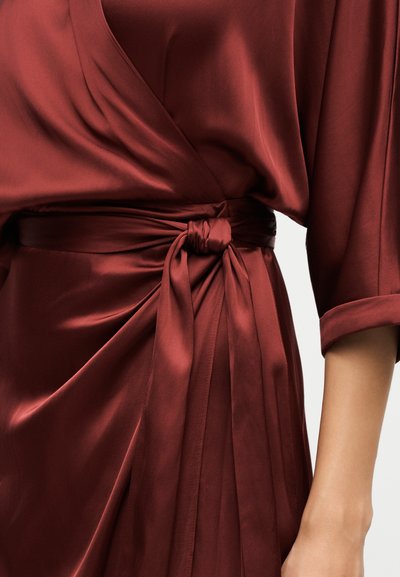 Robe en satin rouge profond avec un design portefeuille, nœud noué à la taille, manches larges aux coudes et texture de tissu lisse et fluide.