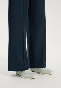 Széles, sötétkék nadrág sima textúrával, világoszöld sportcipővel, lekerekített orral és fehér talppal párosítva.