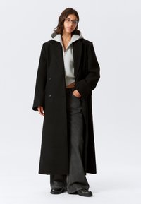 Melns vilnas maisījuma garais mētelis ar dubultrindu pogām, oversized piegriezumu un gludu tekstūru, valkāts pār gaiši pelēku sporta jaku un tumšām džinsām.