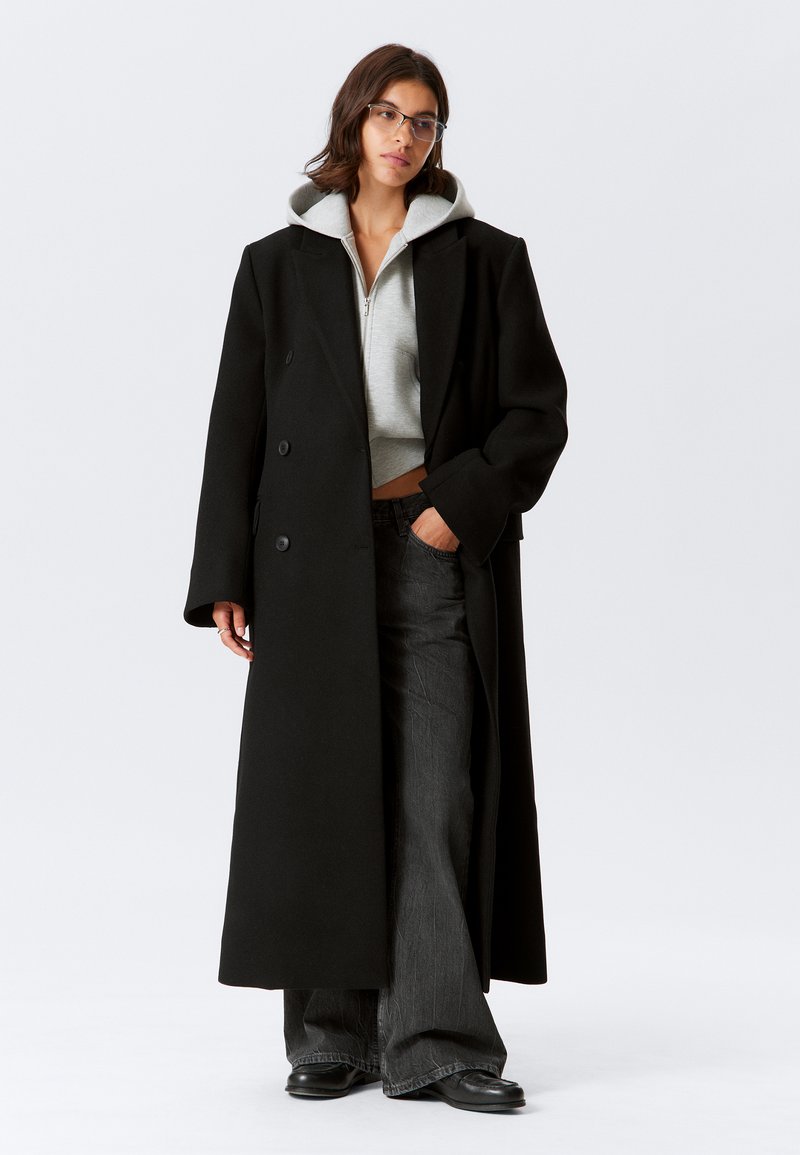 Melns vilnas maisījuma garais mētelis ar dubultrindu pogām, oversized piegriezumu un gludu tekstūru, valkāts pār gaiši pelēku sporta jaku un tumšām džinsām.