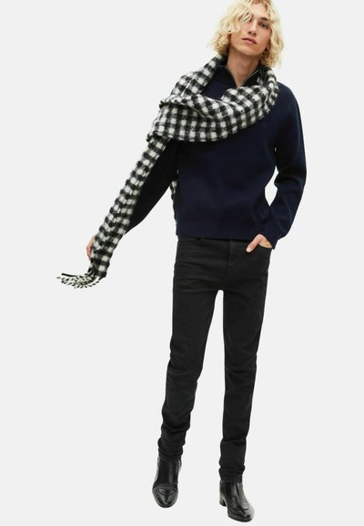 Granatowy sweter z okrągłym dekoltem, czarne jeansy slim fit oraz czarno-biała chusta w kratę zarzucona na ramieniu. Gładka faktura i dopasowany krój.