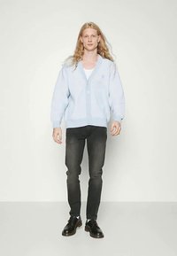 Tânăr cu păr lung și blond, purtând un cardigan butonat de culoare albastră deschisă, cămașă albă, blugi negri și pantofi negri, stând în fața unui perete simplu.