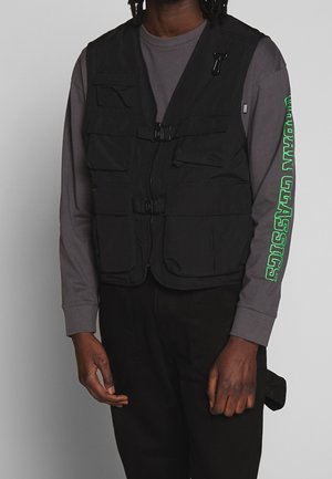 Mann trägt eine schwarze Mehrfach-Taschenweste über einem grauen Langarmshirt mit grünem "Urban Classics"-Text auf dem rechten Ärmel.