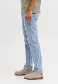 Bărbat purtând blugi albastru deschis, pantofi bej din piele întoarsă cu șireturi și o cămașă verde măsliniu, stând pe un fundal alb.